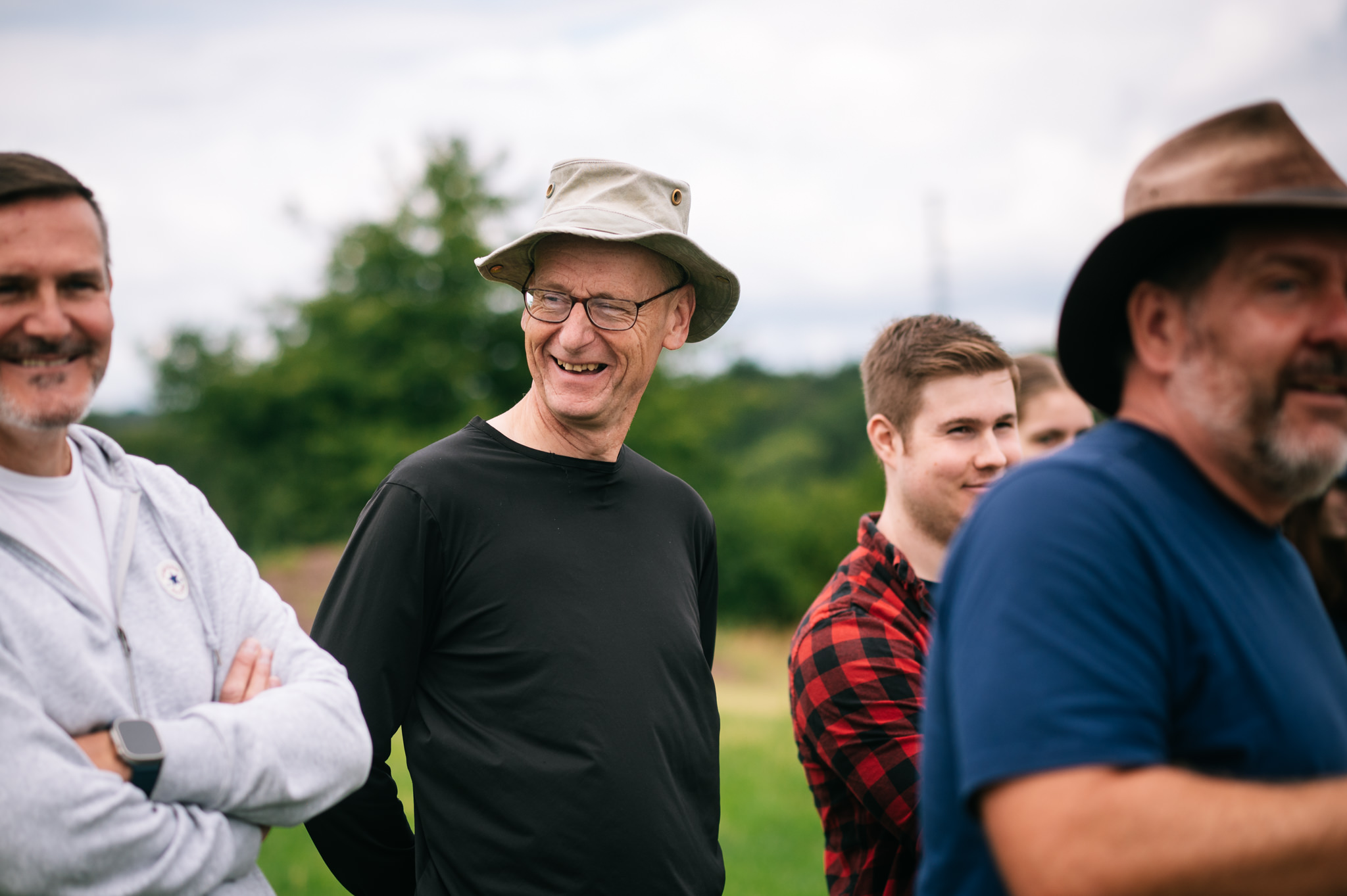 man in a black shirt and light grey hat stood in a field laughing with a group of people