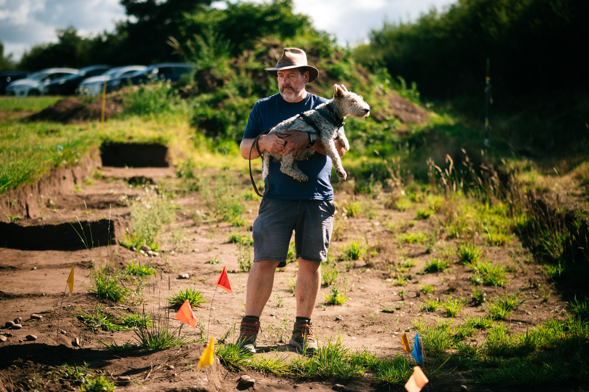 archaeologist in a hat stood in a trench while holding his dog