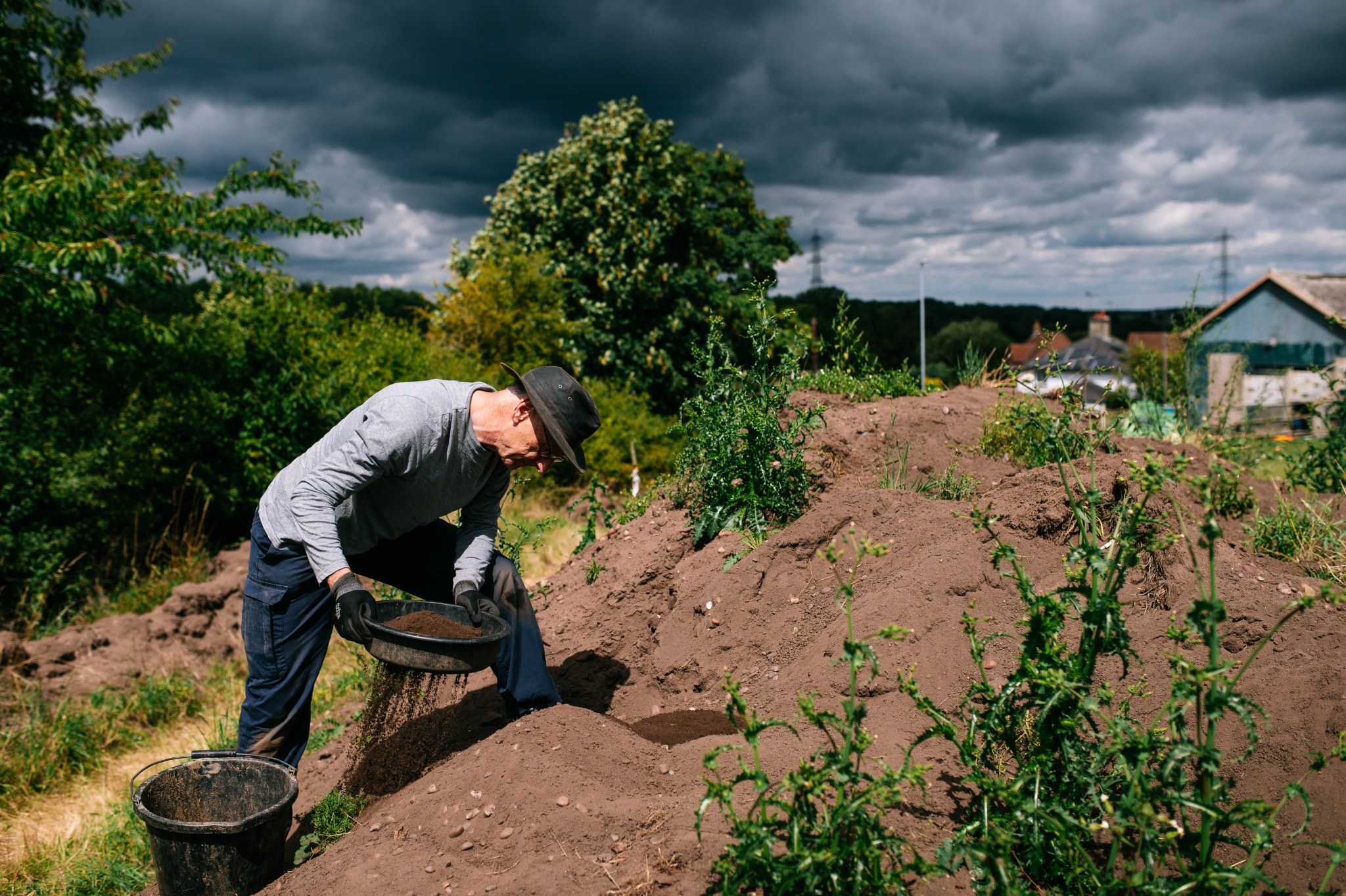 man wearing a hat sieving soil from his trench onto a spoil heap with dark grey clouds behind him