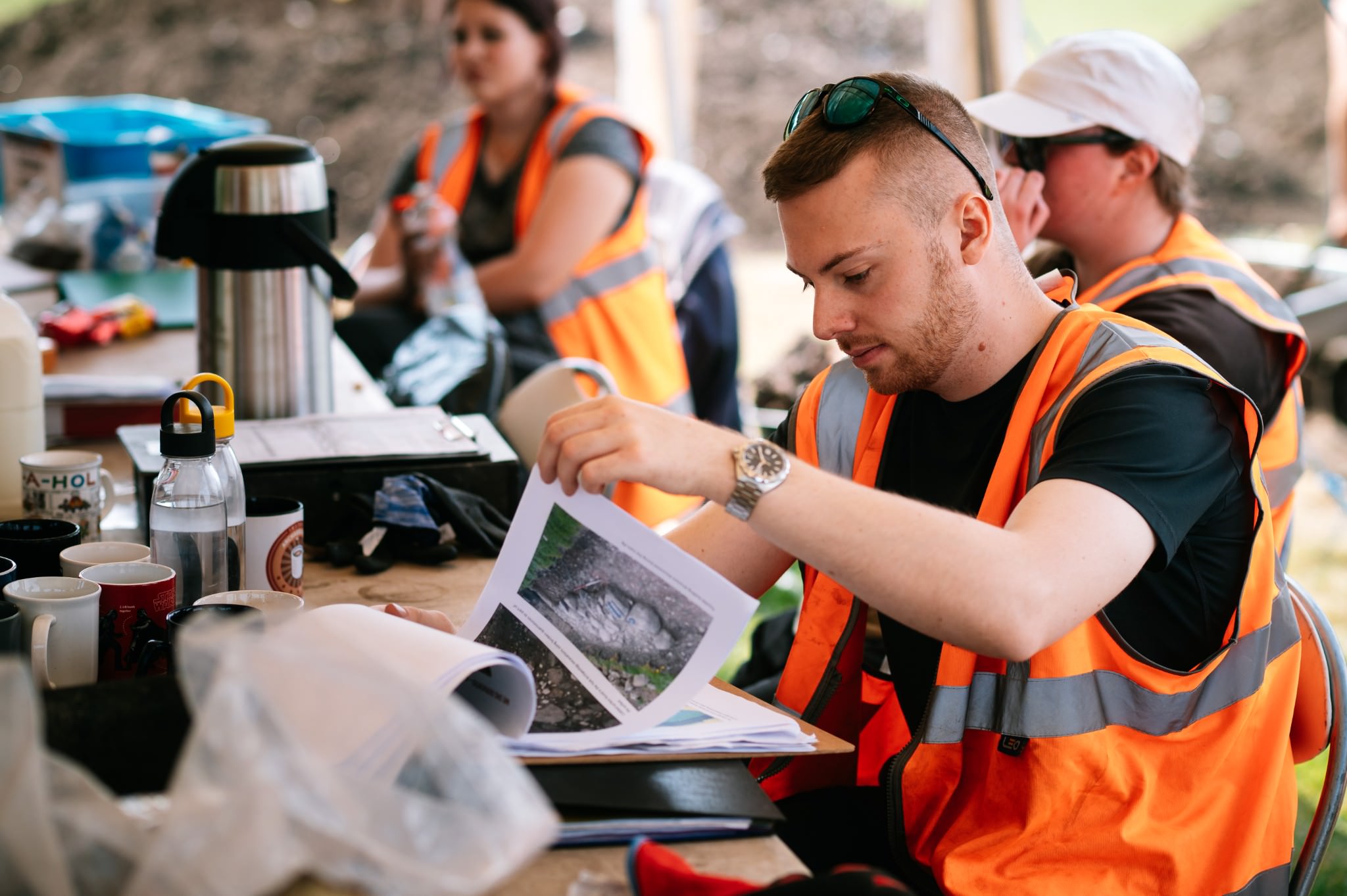 a man in a high vis vest and sunglasses on his head looks through some print outs in an archaeological dig tent